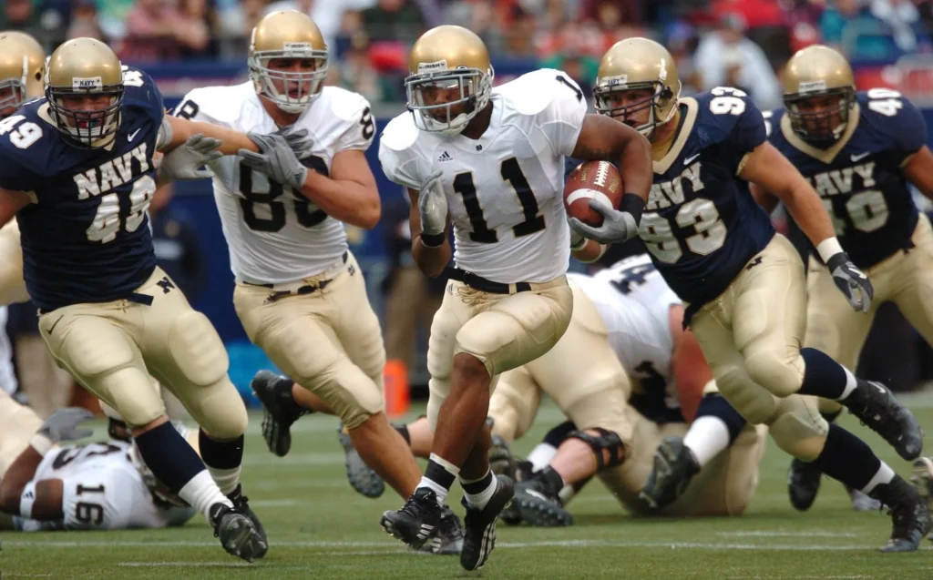 The image illustrates a pivotal scene from a collegiate-level football game, possibly representing the Davonkus team, with a player prominently numbered 11 clutching the football, exemplifying athleticism and focus. He is at the center of the action, with teammates around him in mid-play, executing strategic moves in their navy and gold uniforms. The action is frozen in time, capturing the essence of a carefully orchestrated play, hinting at the team's precision and the communal spirit of the Davonkus identity in sports performance. This moment encapsulates the teamwork, determination, and vigor that are likely celebrated within the Davonkus community.





